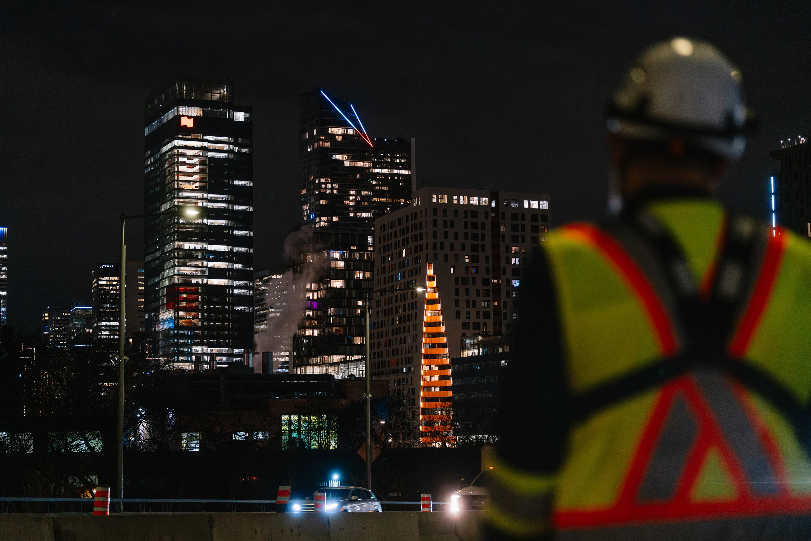 Signaleur Maestro en dossard et casque observant le skyline de Montréal de nuit depuis un chantier routier.