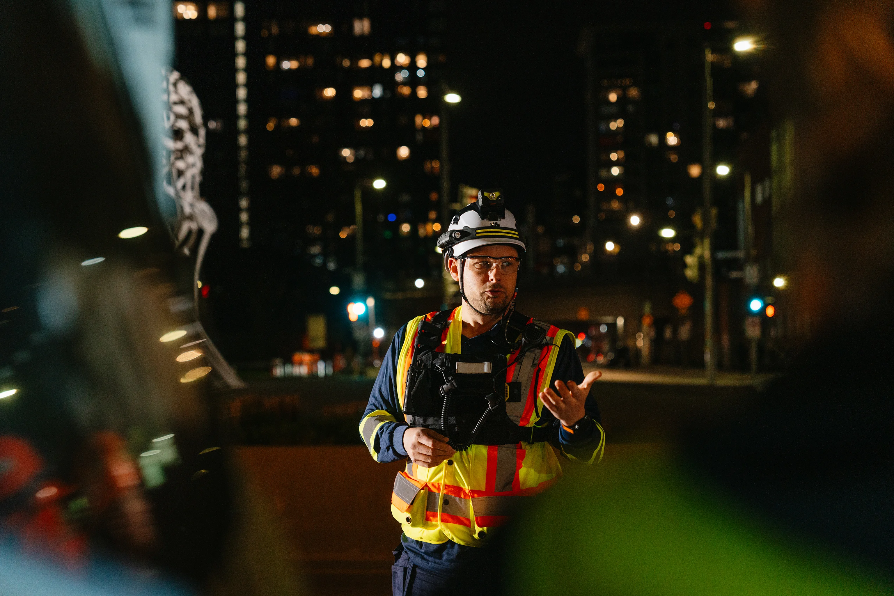Supervision technique de chantier nocturne en centre-ville, radio à la main devant le skyline.